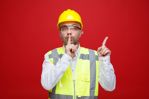 Homme constructeur en uniforme de construction et casque de sécurité portant des lunettes de sécurité regardant la caméra souriant joyeusement faisant un geste silencieux pointant avec l'index sur le côté debout fond rouge