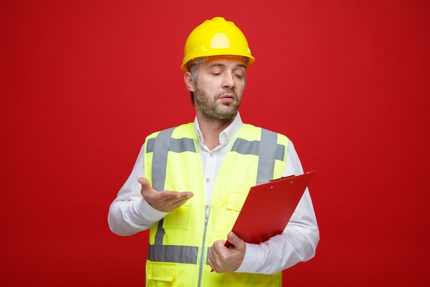 Homme constructeur en uniforme de construction et casque de sécurité tenant le presse-papiers en le regardant confus levant le bras de mécontentement debout sur fond rouge