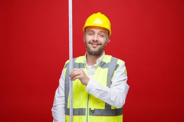 Homme constructeur en uniforme de construction et casque de sécurité tenant un ruban à mesurer regardant la caméra souriant joyeusement heureux et positif debout sur fond rouge