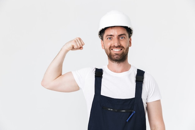 Homme de construction barbu confiant portant une salopette et un casque de sécurité debout isolé sur un mur blanc, fléchissant les muscles