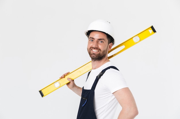 Homme de construction barbu confiant portant une salopette et un casque de sécurité isolé sur un mur blanc, portant un niveau de mesure