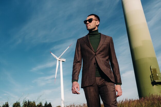 Un homme en costume d'affaires avec une chemise de golf verte se tient à côté d'un moulin à vent sur fond de champ et de ciel bleu. Homme d'affaires près des moulins à vent. Concept moderne du futur.