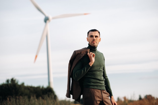 Un homme en costume d'affaires avec une chemise de golf verte se tient à côté d'un moulin à vent sur fond de champ et de ciel bleu. Homme d'affaires près des moulins à vent. Concept moderne du futur.