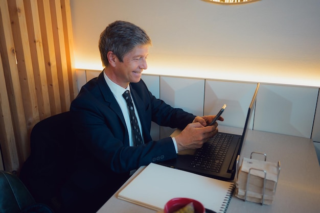 Un homme en costume assis à table avec un ordinateur portable qui regarde son téléphone portable