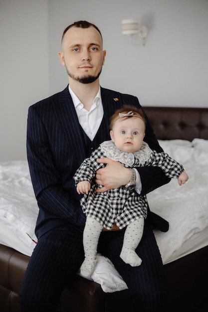 Un homme en costume avec un bébé sur ses genoux est assis sur un lit.