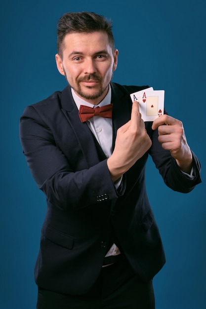 Homme en costume classique noir et noeud papillon rouge montrant deux cartes à jouer tout en posant contre un studio bleu ...
