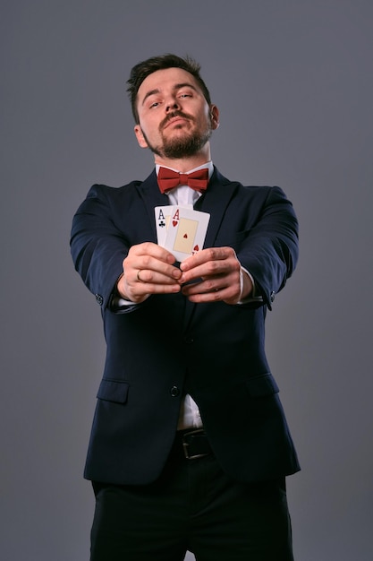 Homme en costume classique noir et noeud papillon rouge montrant deux cartes à jouer tout en posant contre un studio gris ...