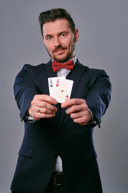 Homme en costume classique noir et noeud papillon rouge montrant deux cartes à jouer tout en posant contre un studio gris ...