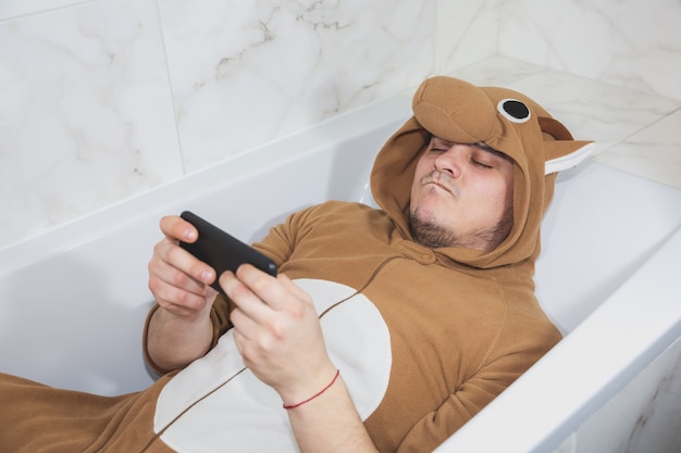 Homme en costume cosplay d'une vache. Un gars en pyjama d'animaux amusants est allongé dans le bain et joue à des jeux sur son téléphone portable.