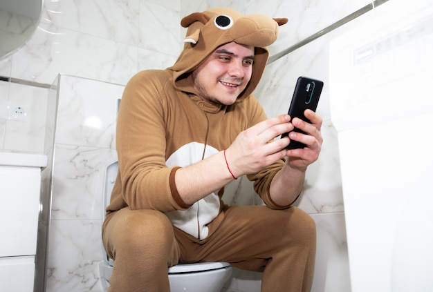 Homme en costume cosplay d'une vache. Un gars en pyjama d'animaux amusants s'assoit sur les toilettes et joue à des jeux sur son téléphone portable.