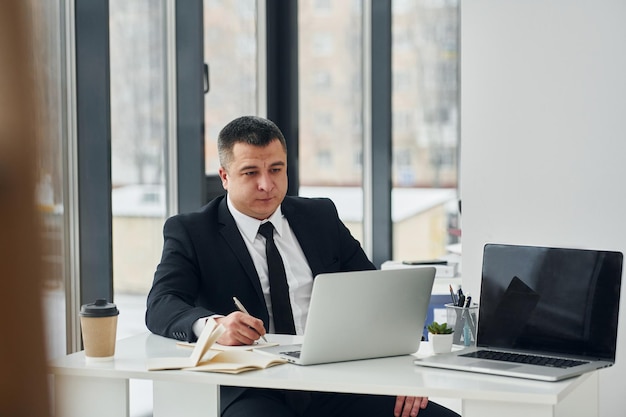 L'homme en costume et cravate est assis à table avec un ordinateur portable et travaille au bureau