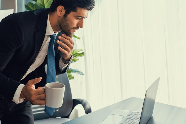 Un homme en costume est assis à un bureau avec un ordinateur portable et regarde un ordinateur portable.