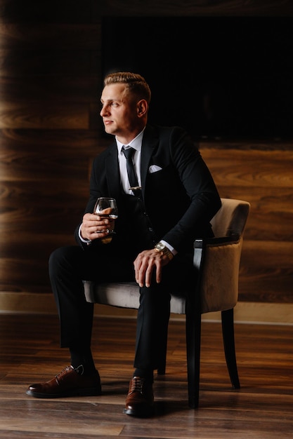 un homme en costume est assis sur une chaise avec un verre de vin