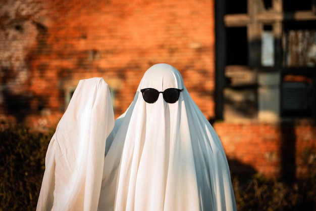 Un homme en costume de fantôme fait d'un drap et de lunettes de soleil se tient près d'un bâtiment abandonné et agite la main Défi fantôme 2021 Saison effrayante Célébration d'Halloween