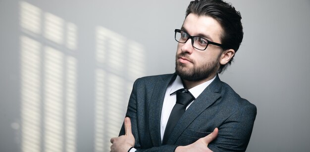 Un homme en costume et lunettes se tient les bras croisés sur un mur gris