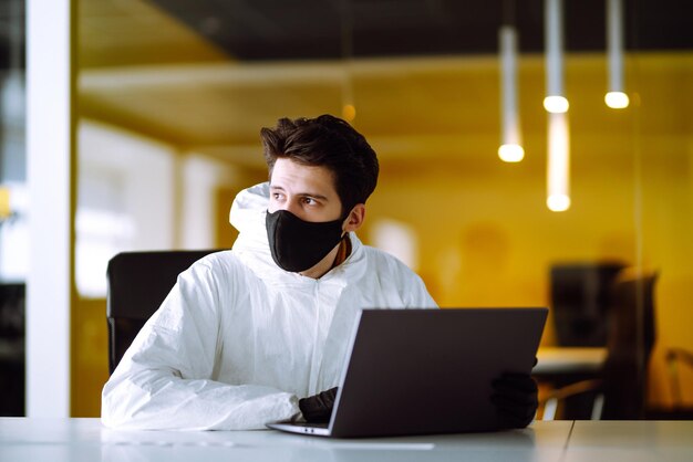 Un homme en costume et masque de protection contre les matières dangereuses travaille sur un ordinateur dans un bureau vide.