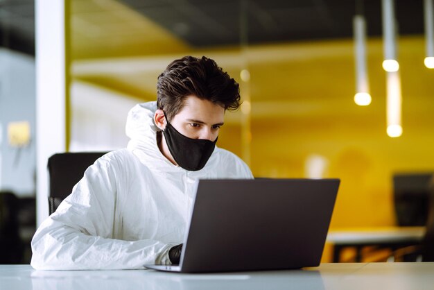 Un homme en costume et masque de protection contre les matières dangereuses travaille sur un ordinateur dans un bureau vide.