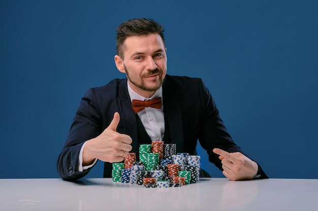 L'homme En Costume Noir Est Assis à Une Table Blanche Avec Des Piles Colorées De Jetons Dessus Posant Sur Un Studio Bleu...