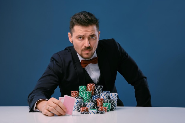 L'homme en costume noir est assis à une table blanche avec des piles de jetons tenant deux cartes à jouer posant sur...