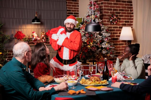 Homme en costume de père noël sac de transport avec des cadeaux, famille échangeant des cadeaux de noël. Diverses personnes célébrant Noël à la maison, mangeant un repas traditionnel à une table de fête