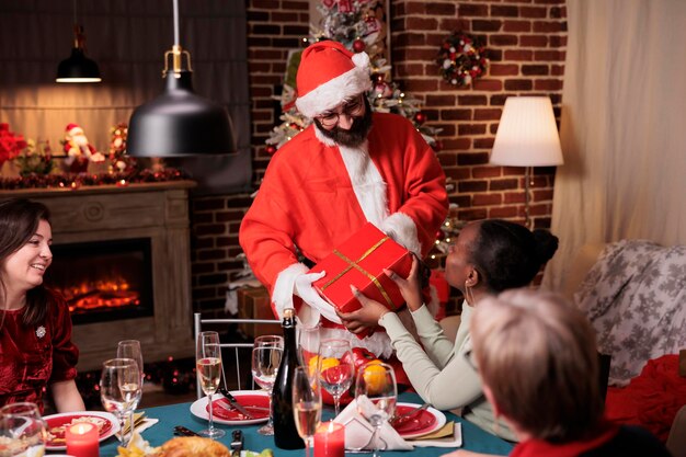 Homme en costume de père noël saluant une femme afro-américaine, donnant un cadeau de noël, parents échangeant des cadeaux à la table de fête. Célébration de la veille de Noël, famille diversifiée mangeant un repas traditionnel