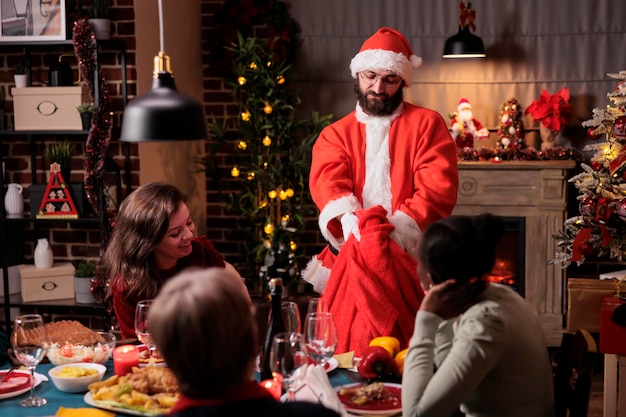 Homme en costume de père noël sortant des cadeaux du sac rouge, grande famille heureuse échangeant des cadeaux de noël. Diverses personnes célébrant Noël à la maison, mangeant un repas traditionnel à une table de fête