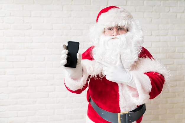 Un homme en costume de Père Noël tenant un téléphone portable contre un mur de briques