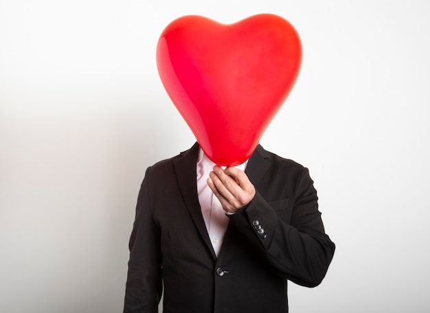 Homme en costume se cachant derrière un ballon en forme de coeur rouge. Homme tenant le symbole de l'amour, de la famille, de l'espoir.