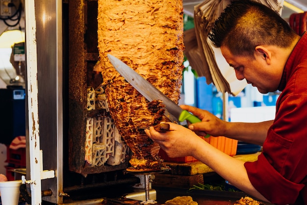 Homme coupant des morceaux d'un morceau de viande dans une toupie pour tacos al pastor, plat typiquement mexicain.