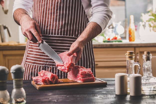 L'homme Coupe Un Morceau De Viande Fraîche Sur Une Planche à Découper En Bois Dans La Cuisine à Domicile. Un Homme Dans Un Tablier Rayé Avec Un Grand Couteau Dans Ses Mains