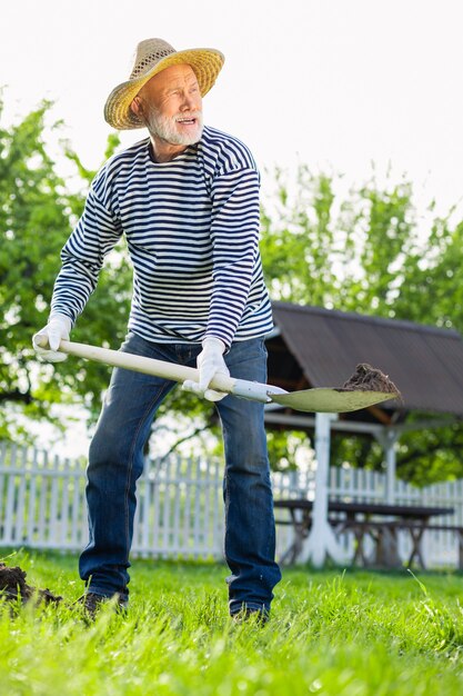 Homme creusant le sol. Homme aux cheveux gris en chapeau de paille et chemise rayée à l'aide d'une pelle tout en creusant le sol