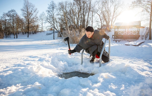 Un homme creuse un trou dans la glace avec une hache pour nager en hiver dans une journée d'hiver froide et ensoleillée