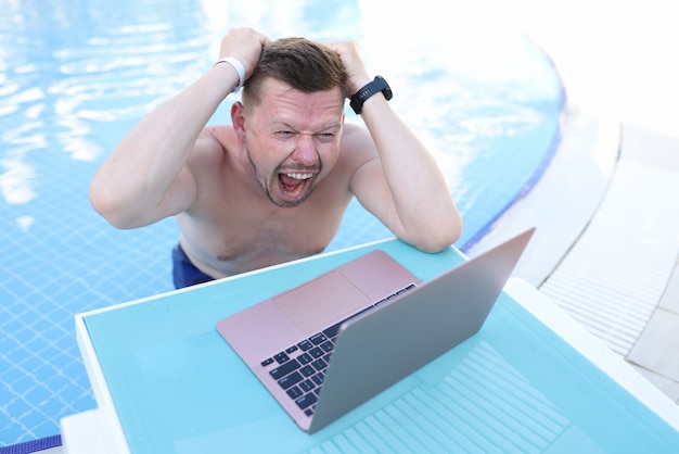 Homme criant et tenant ses cheveux et regardant un écran d'ordinateur portable sur le côté de la piscine