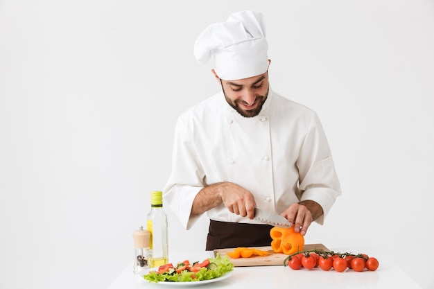 homme cuisinier caucasien en uniforme souriant et coupant une salade de légumes sur une planche de bois isolée sur un mur blanc