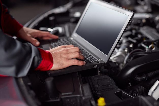 Homme de culture utilisant un netbook avec écran noir tout en vérifiant le moteur d'un véhicule moderne pendant le travail dans le garage