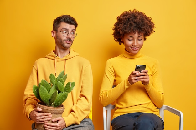 Un homme curieux jette un coup d'œil dans le smartphone de sa petite amie essaie de voir le contenu du message vêtu d'un sweat-shirt contient un pot de cactus.