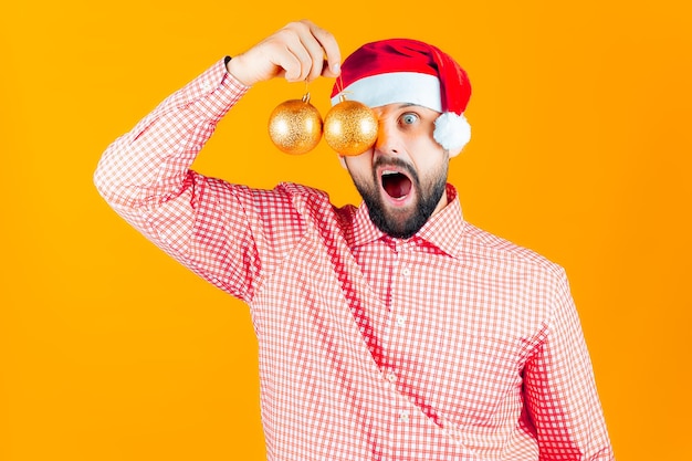 Photo un homme dans un chapeau de noël du père noël tient devant ses yeux des jouets du nouvel an, des boules de couleur dorée et des cris.