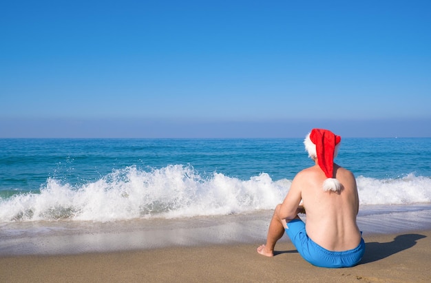 Un homme dans un chapeau de Père Noël est assis sur la vue arrière du bord de mer