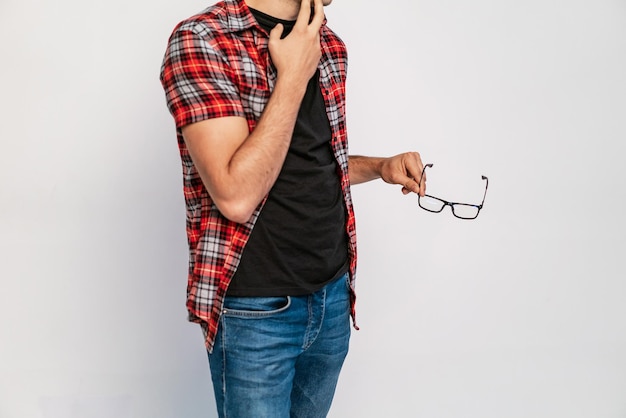 Un homme dans une chemise à carreaux rouge et un jean tient le temple de lunettes par sa main sur un fond blanc Gros plan