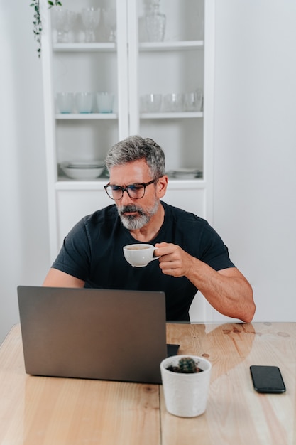 Homme dans la cinquantaine travaillant à domicile tout en prenant un café et est en appel vidéo sur l'ordinateur