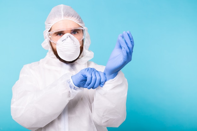 Homme dans un costume de décontamination blanc mettant des gants médicaux isolés sur un mur bleu