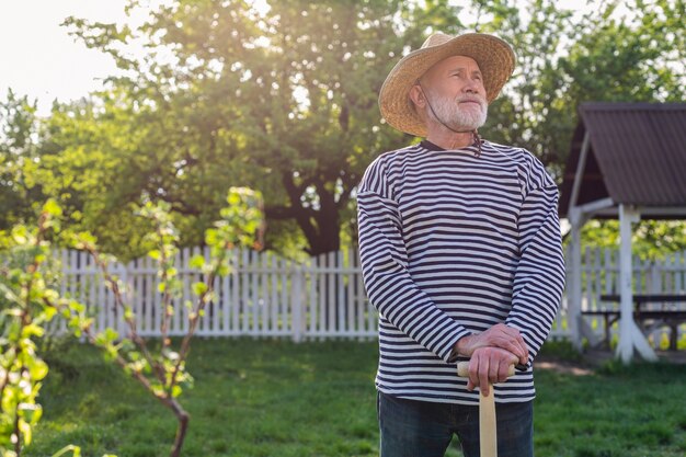 L'homme dans le jardin. Homme à la retraite aux cheveux gris portant un chapeau de paille tenant une pelle debout dans son jardin