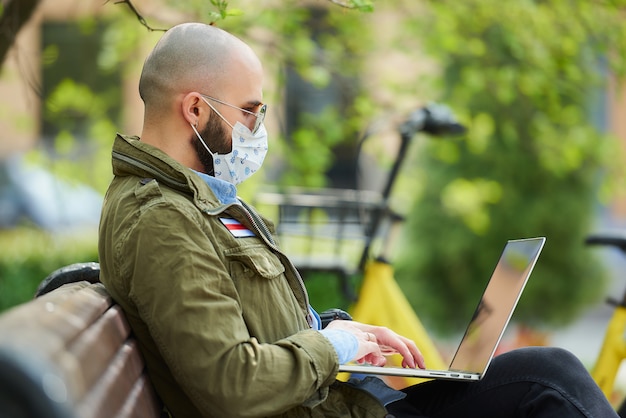 Un homme dans un masque facial pour éviter la propagation du coronavirus travaille à distance sur un ordinateur portable dans le parc. Un gars porte des lunettes de soleil est assis sur un banc dans la rue avec un ordinateur près d'un vélo. Drapeau blanc-rouge-blanc.