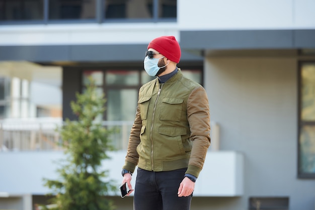 Un homme dans un masque médical pour éviter la propagation du coronavirus tenant son smartphone dans une rue confortable.