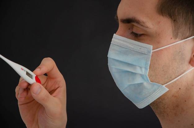 Un homme dans un masque médical tient, regarde un thermomètre.