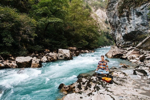 Photo un homme dans une position méditative avec une guitare assis sur la rive d'une rivière de montagne sur un fond de