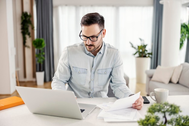 Un homme dans sa maison confortable regardant l'ordinateur portable travaillant sur la paperasse et résolvant les finances de la maison