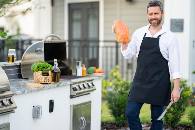 L'homme Dans Le Tablier Du Chef Tient Du Poisson Saumon Au Barbecue Grill Cuisinier Mâle Préparant Un Barbecue à L'extérieur Du Poisson Barbecue