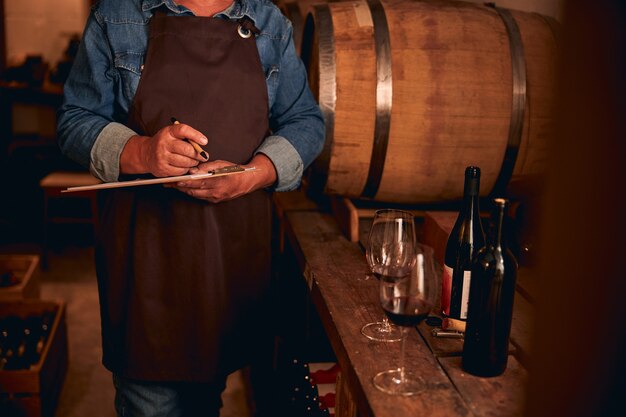 homme dans un tablier marron debout avec un presse-papiers et un stylo à côté d'un tonneau de vin. Bouteilles et verres sur la table