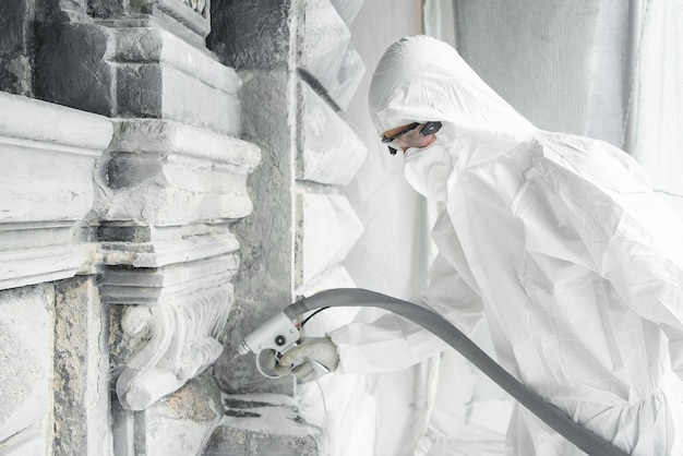 L'homme dans un uniforme de protection blanc nettoie la sculpture en pierre sculptée de la saleté et du béton avec une machine de sablage. Restauration de la sculpture sur pierre. Un jet de sable sous haute pression.